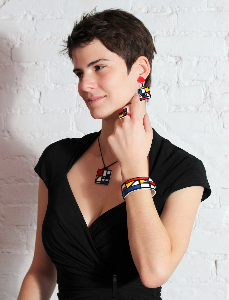 A model wearing the Riemondrian jewelry set of laser-cut acrylic earrings, ring, pendant necklace, and bracelet, each with a Mondrian pattern of red, yellow, blue, and white blocks with black lines